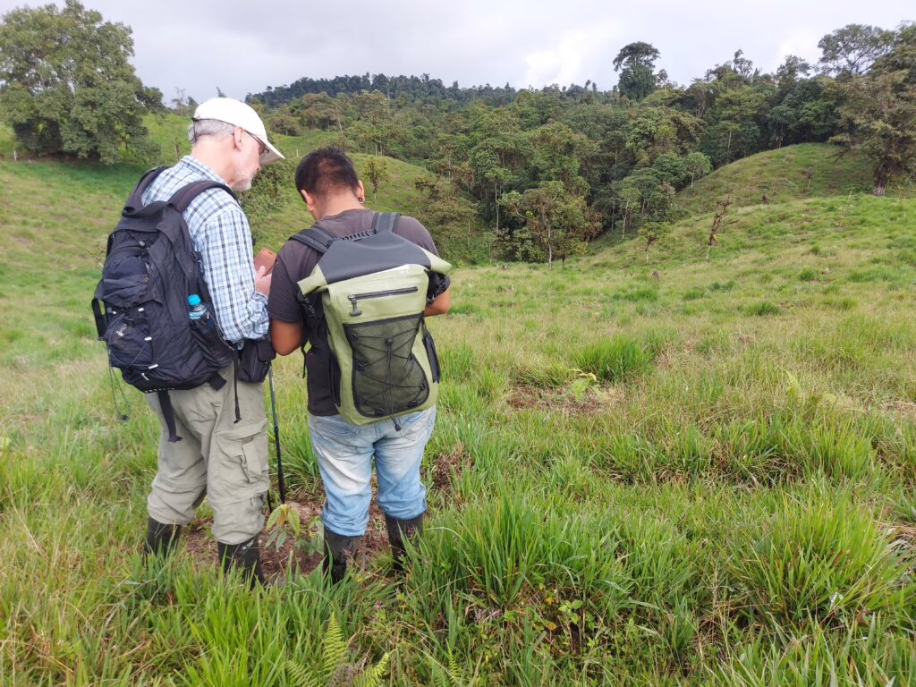 Foto wurde auf einer grünen Wiese aufgenommen. Zu sehen sind zwei Menschen von hinten, die sich etwas anschauen. Weit hinten ist großer Wald.