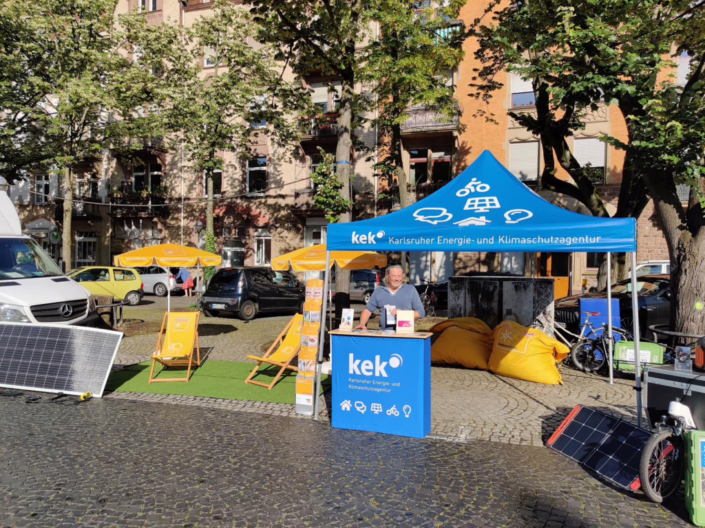 Mitarbeiter Christian Diebold steht beim Infostand der KEK auf dem Gutenbergplatz und lächelt in die Kamera. Im Hintergrund ist eine Hausfassade und große Bäume. 