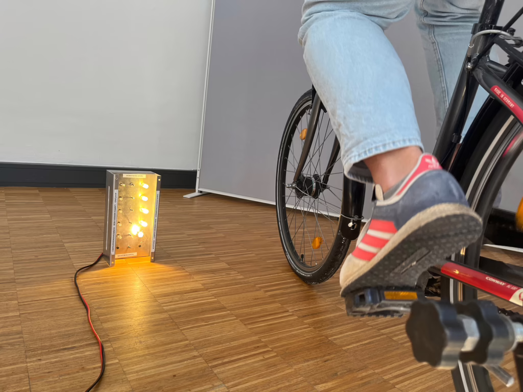 Eine Person fährt auf einem Fahrrad das in eine Maschine eingespannt ist. Dadurch fährt es nicht, sondern bringt Lampen zum leuchten. 