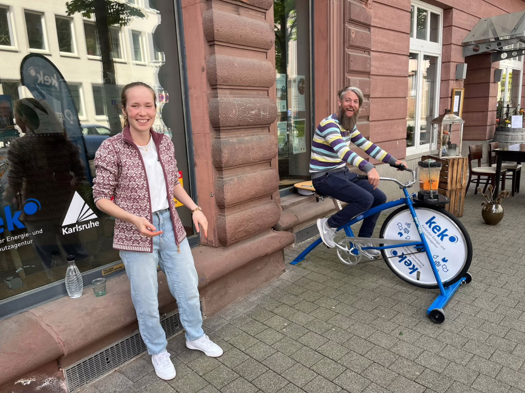 Zwei Mitarbeiterinnen und Mitarbeiter der KEK sind vor dem Beratungszentrum. Beide lächeln in die Kamera. Ein mitarbeiter sitzt auf einem Fahrrad, das nicht fährt, sondern mit dem man eine Smoothie-Maschine betreiben kann. 