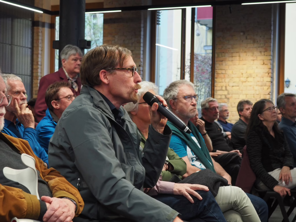 Ein Mann sitzt im Publikum und stellt eine Frage an die Referenten und spricht in ein Mikrofon.